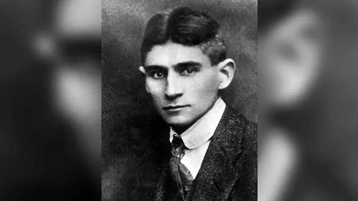 Un portrait en noir et blanc d'un jeune homme aux cheveux sombres coupés court,  vêtu d'une veste en tweed et d'une cravate.