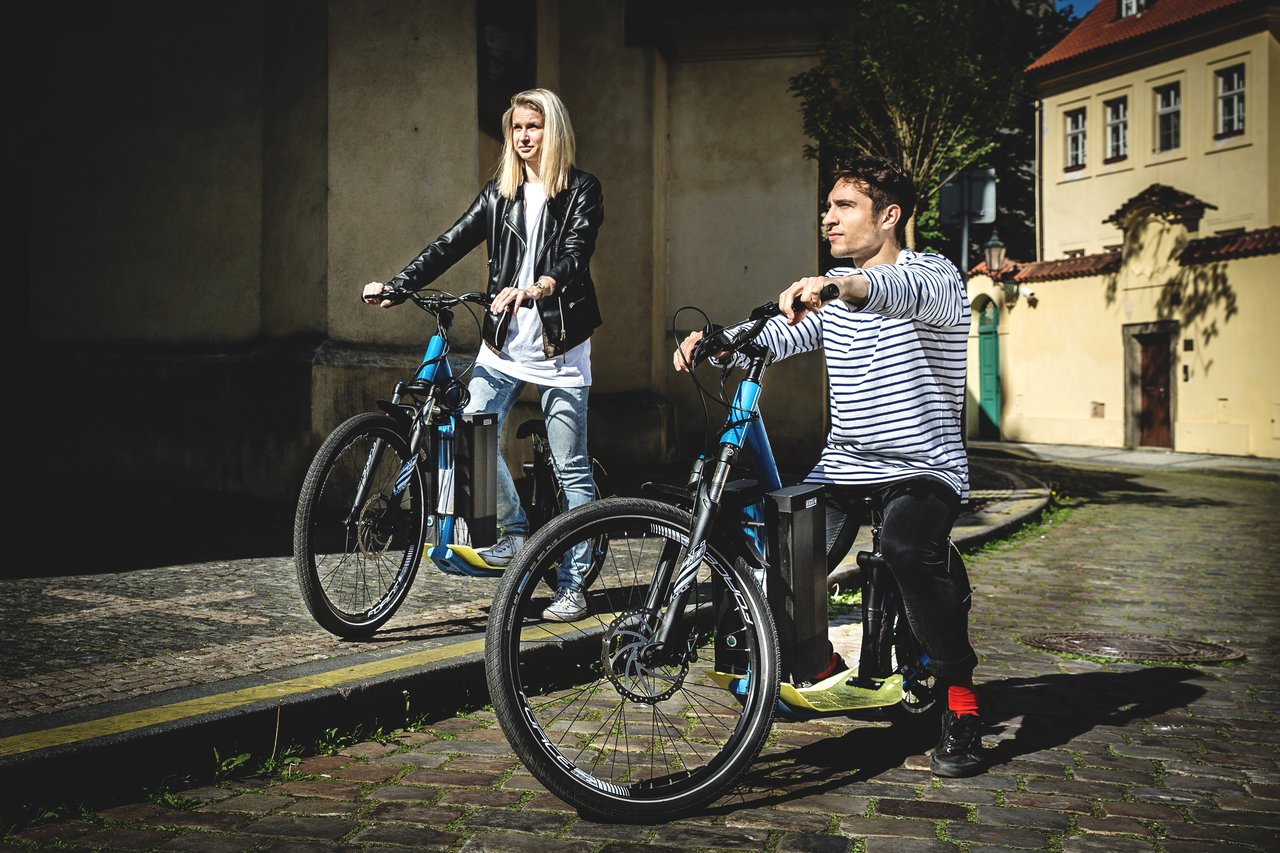 Deux personnes sur une trottinette électrique, le nez rivé vers l'horizon, roulant sur les rues pavées de Prague