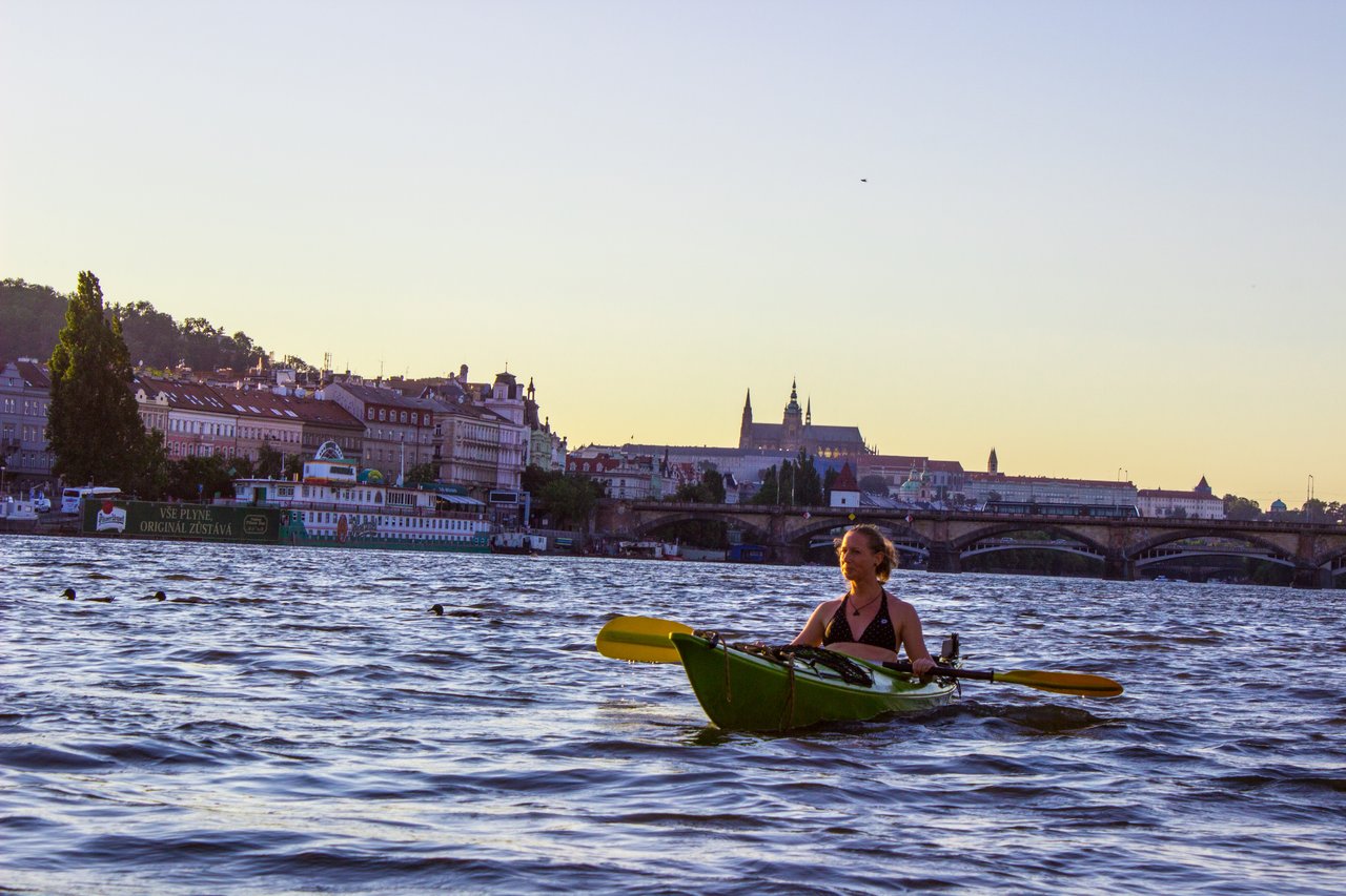 Une dame qui fait du kayak à gauche on peut voir des canards qui nagent et en fond il y a la ville