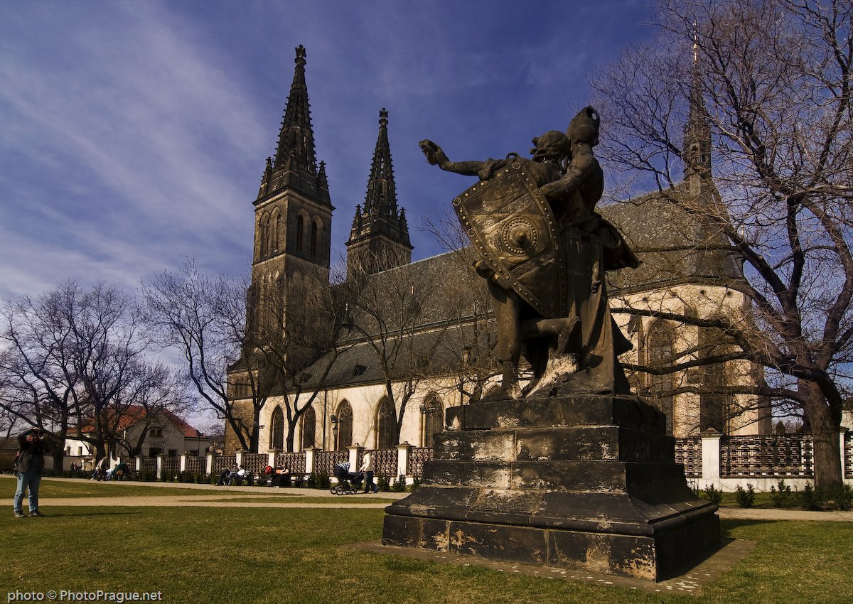 Statue en pierre d'un guerrier portant un bouclier au milieu d'un parc, devant une église dotée de hautes flèches noires