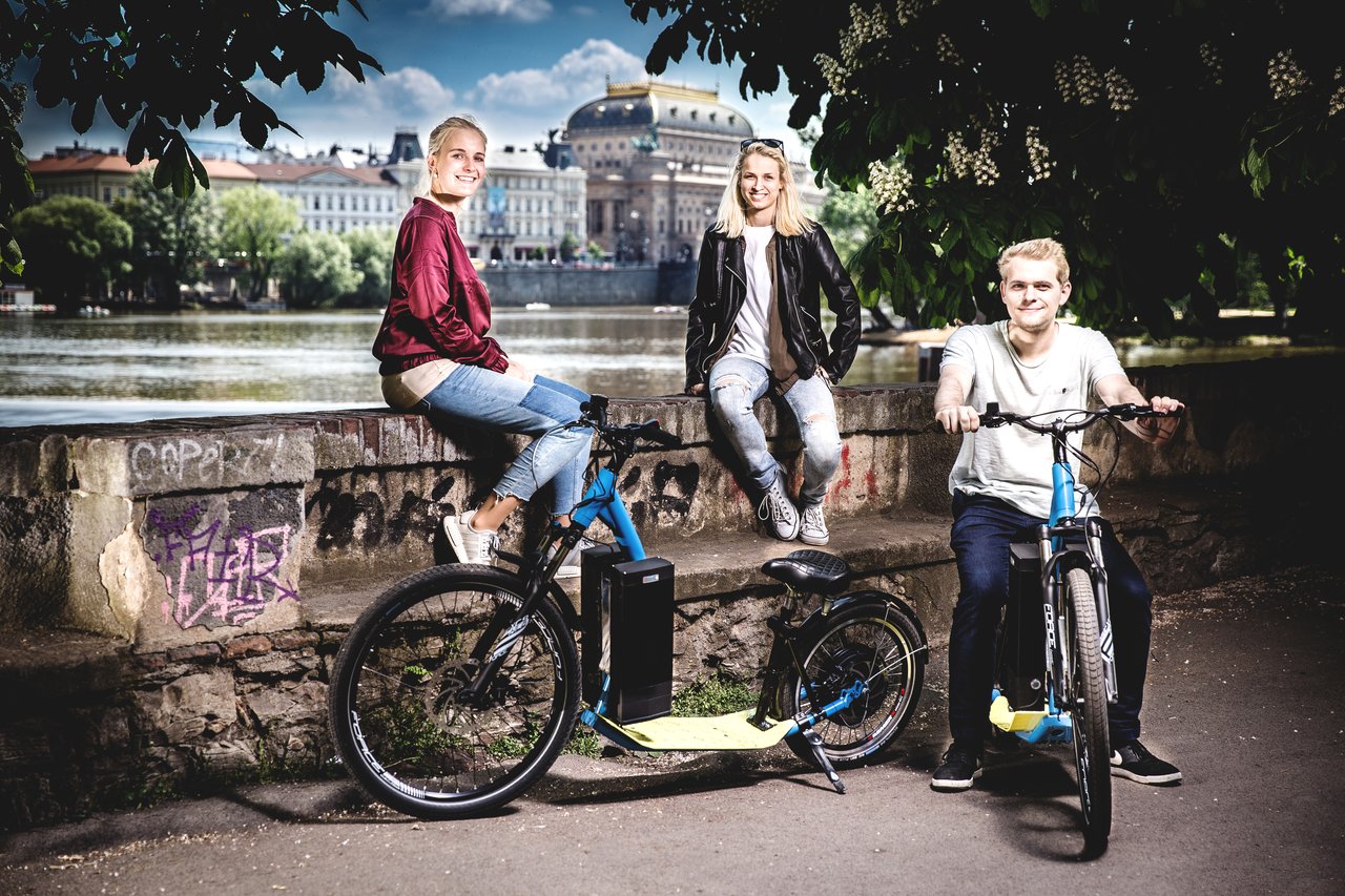 Deux personnes assises sur un mur aux abords de Prague, avec un homme assis sur un scooter électrique devant elles