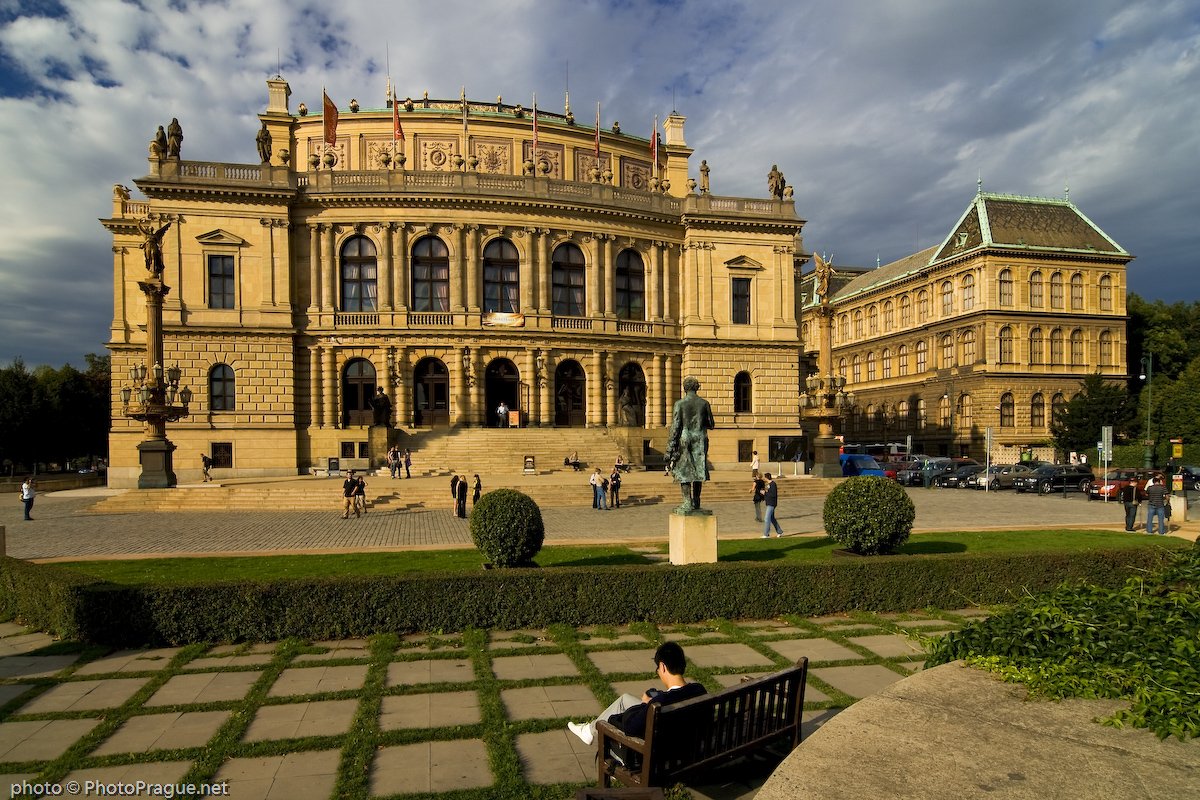 Une imposante façade de bâtiment en pierre beige,ornée de colonnes et de statues,avec devant une place la statue