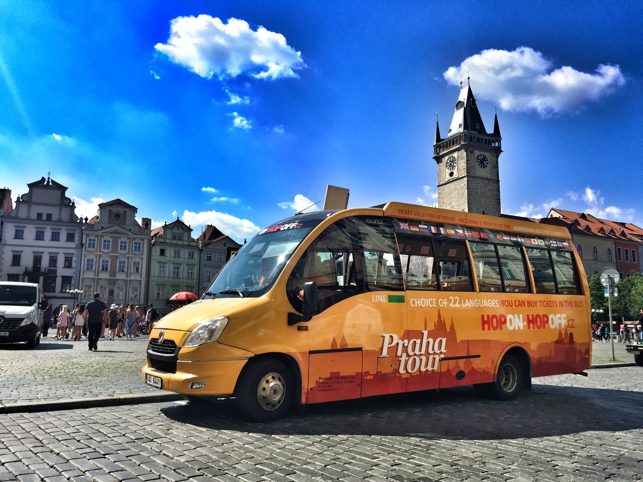 Un bus touristique orange et jaune est stationné sur une place ensoleillée, devant des bâtiments anciens et une haute tour d'horloge