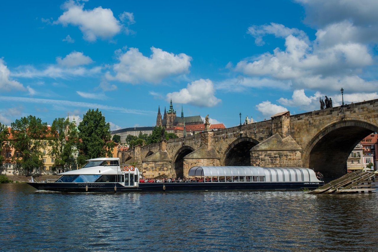 Grand bateau qui traverse sous un pont avec le toit à moitié ouvert, il y a des gens dedans en font on peut voir la ville
