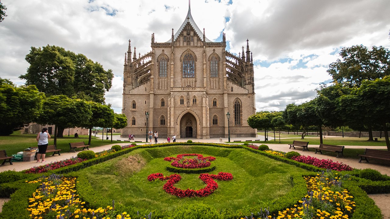 Une grande église en pierre aux toits très pointus s'élève derrière un jardin d'une pelouse verte et de fleurs coloré