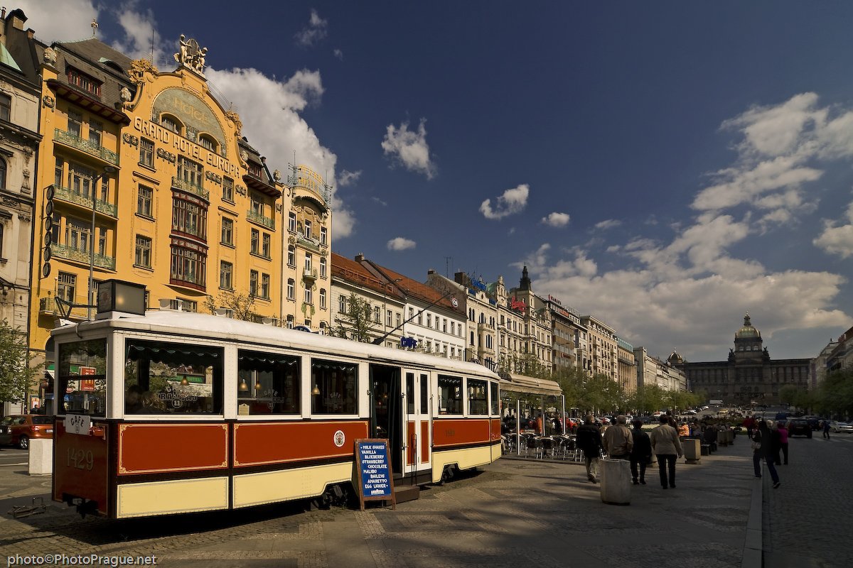 Un vieux tramway rouge et blanc est garé sur une place  bordée de grands bâtiments colorés, sous un ciel bleu parsemé de nuage