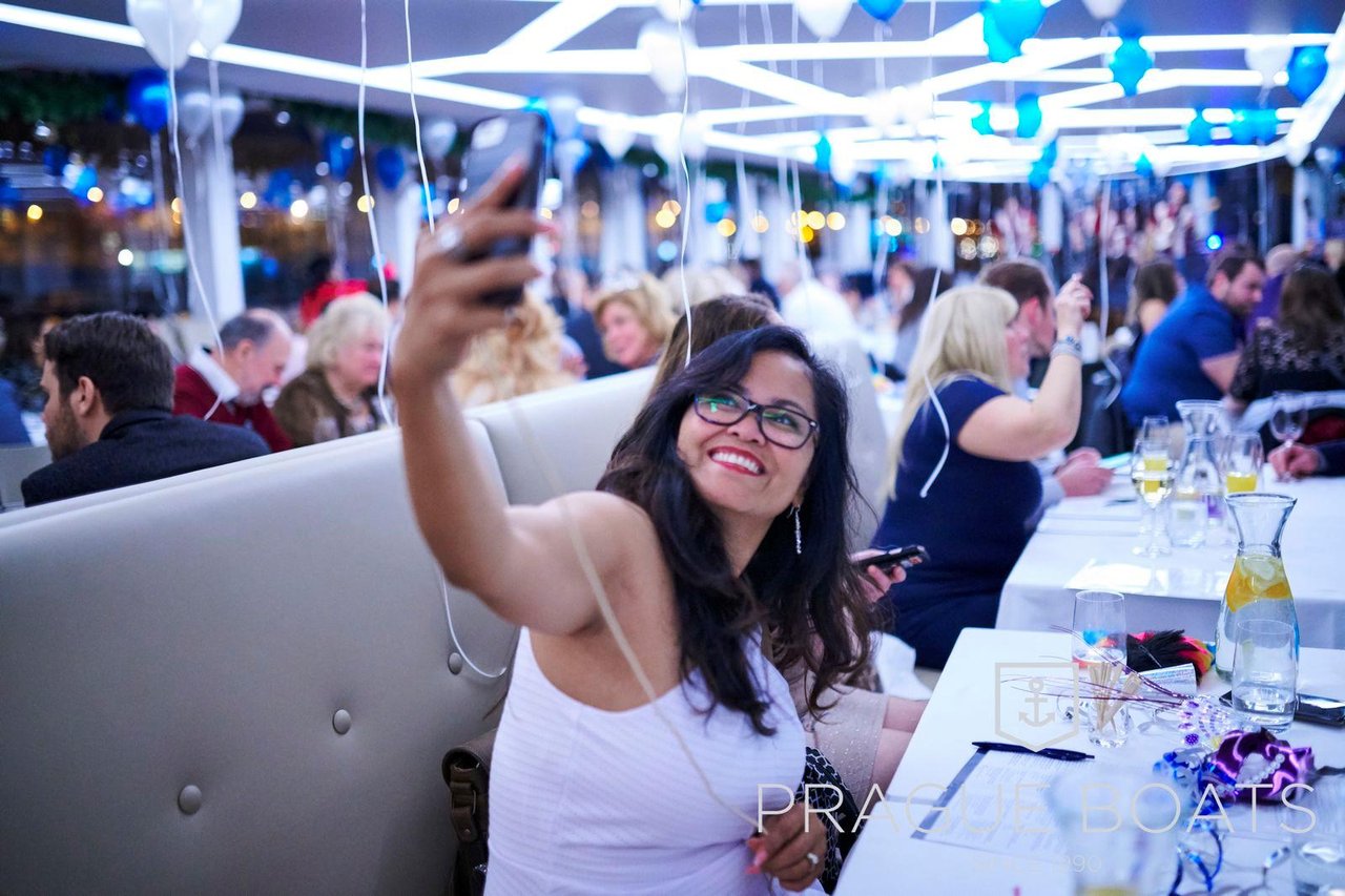 Une femme qui prend un selfie dans une foule assis sur des chaises, au-dessus il y a des ballons bleu et blanc