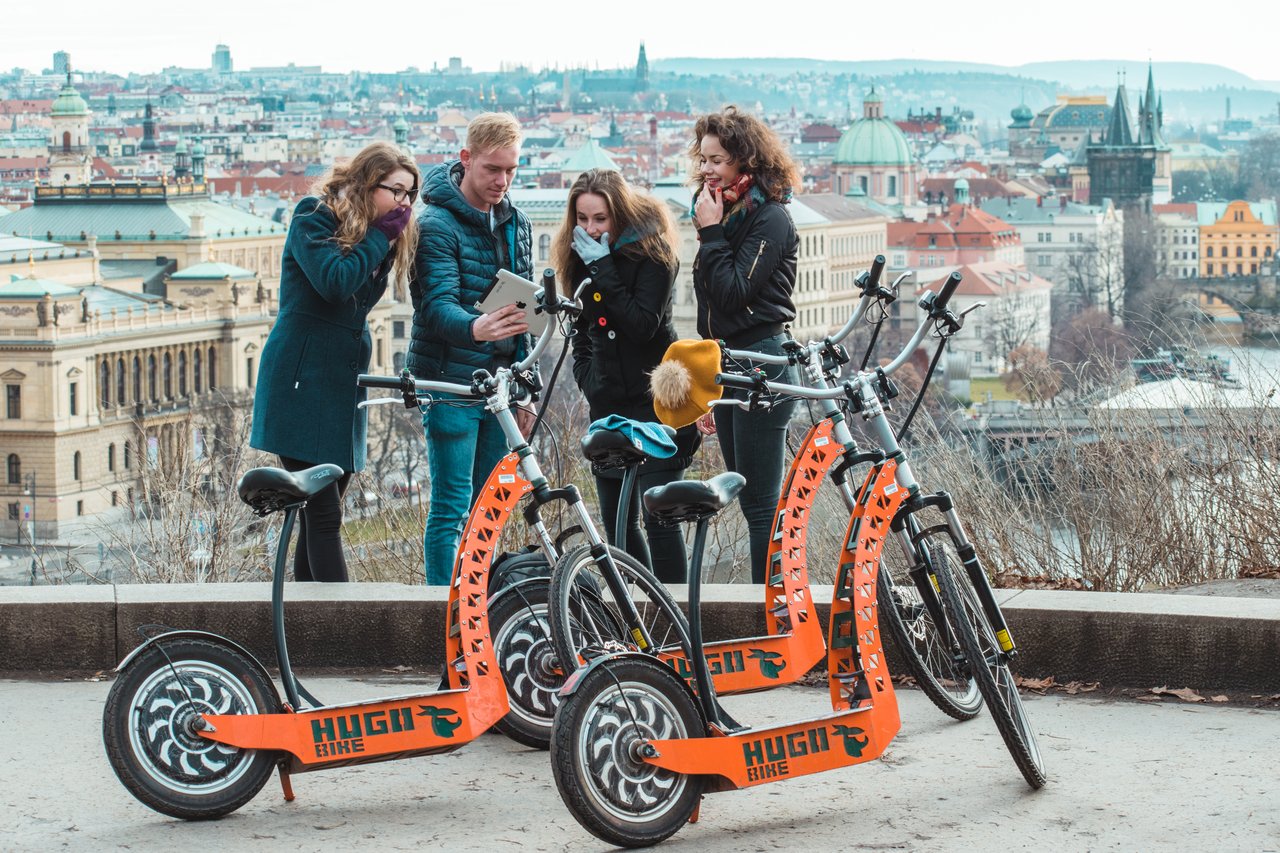 4 personnes regardant une tablette avec un air surpris, debout devant  3 trottinettes électriques, avec une vue sur la ville