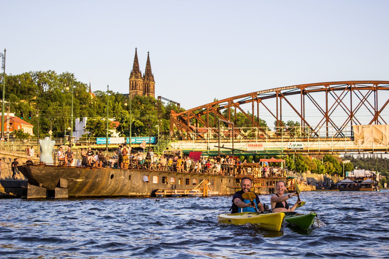Un homme et une femme qui font du kayak en fond il y a un vieux bateau avec des gens dessus et on peut voir le pont