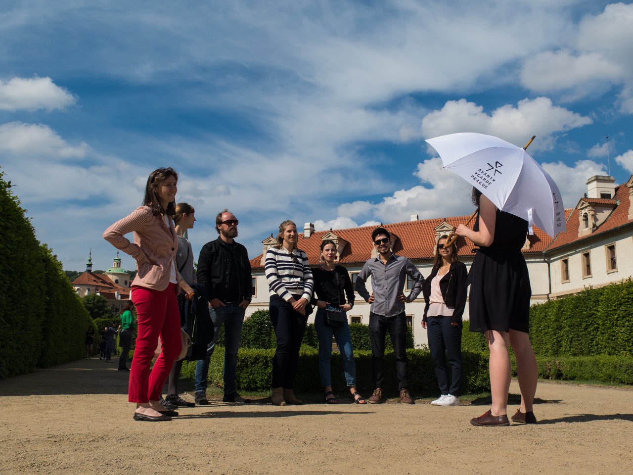 Un guide touristique, un parapluie à la main,s'adresse à sept personnes dans un magnifique jardin,sous un ciel ensoleillé.