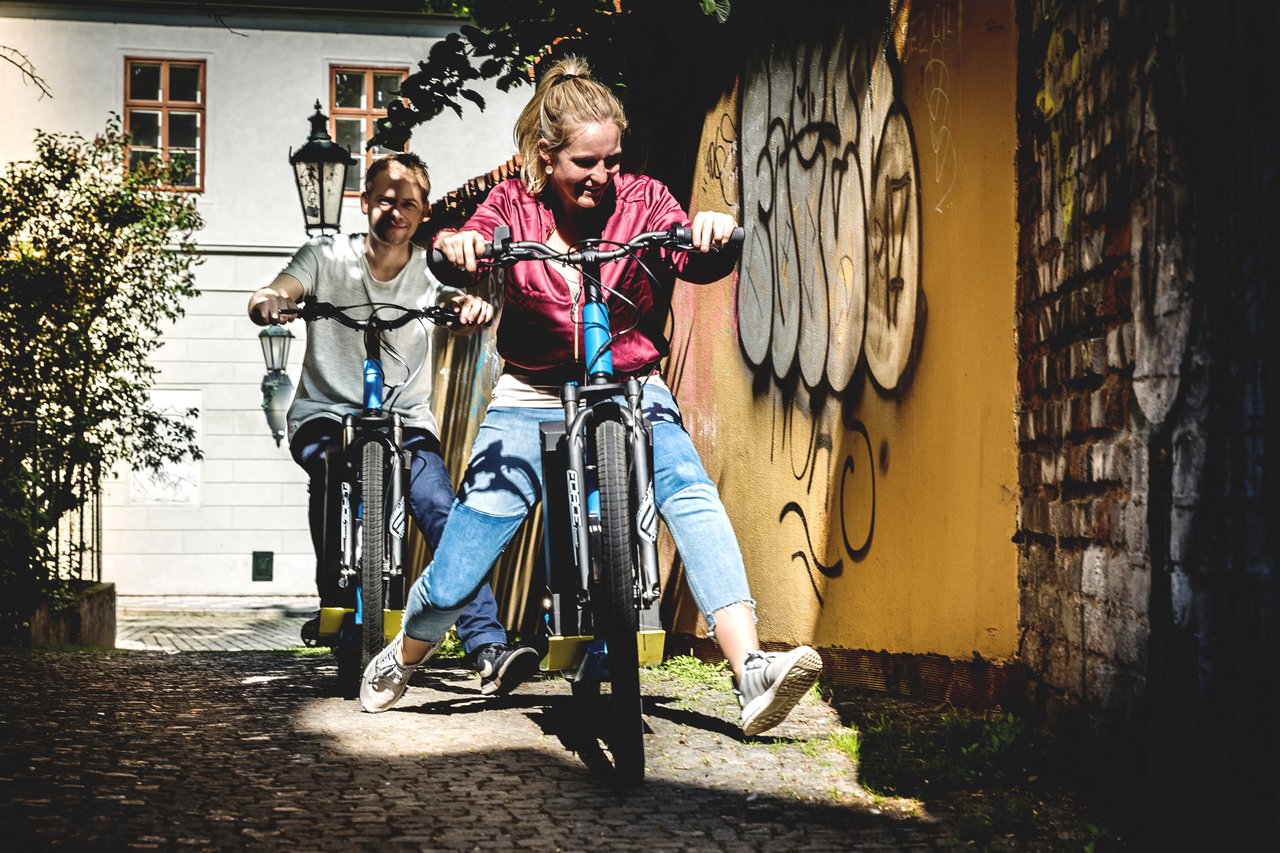 Deux personnes souriantes sur une trottinette électrique qui descendent une petite rue bordée de murs en pierre