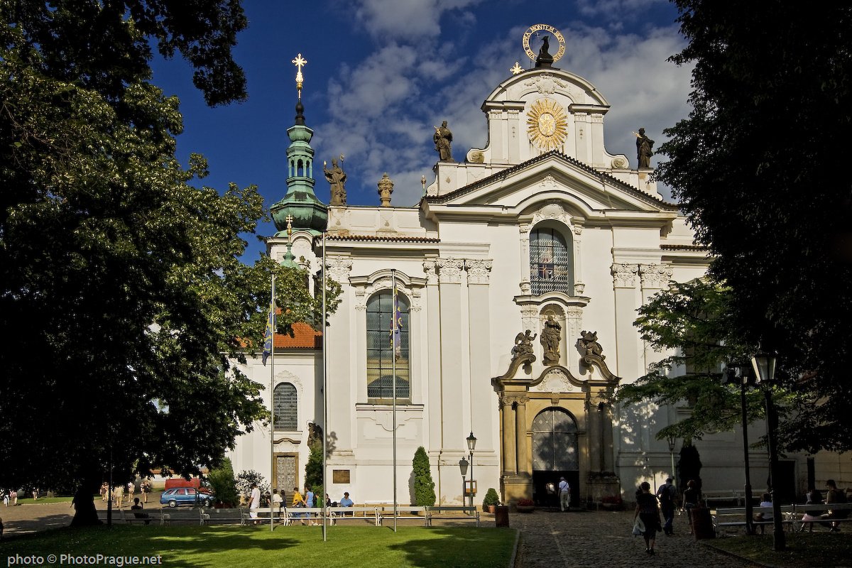 Une façade blanche ornée d'une statue et de moulures, entourée d'arbres et de passants sous un ciel bleu et nuageux.
