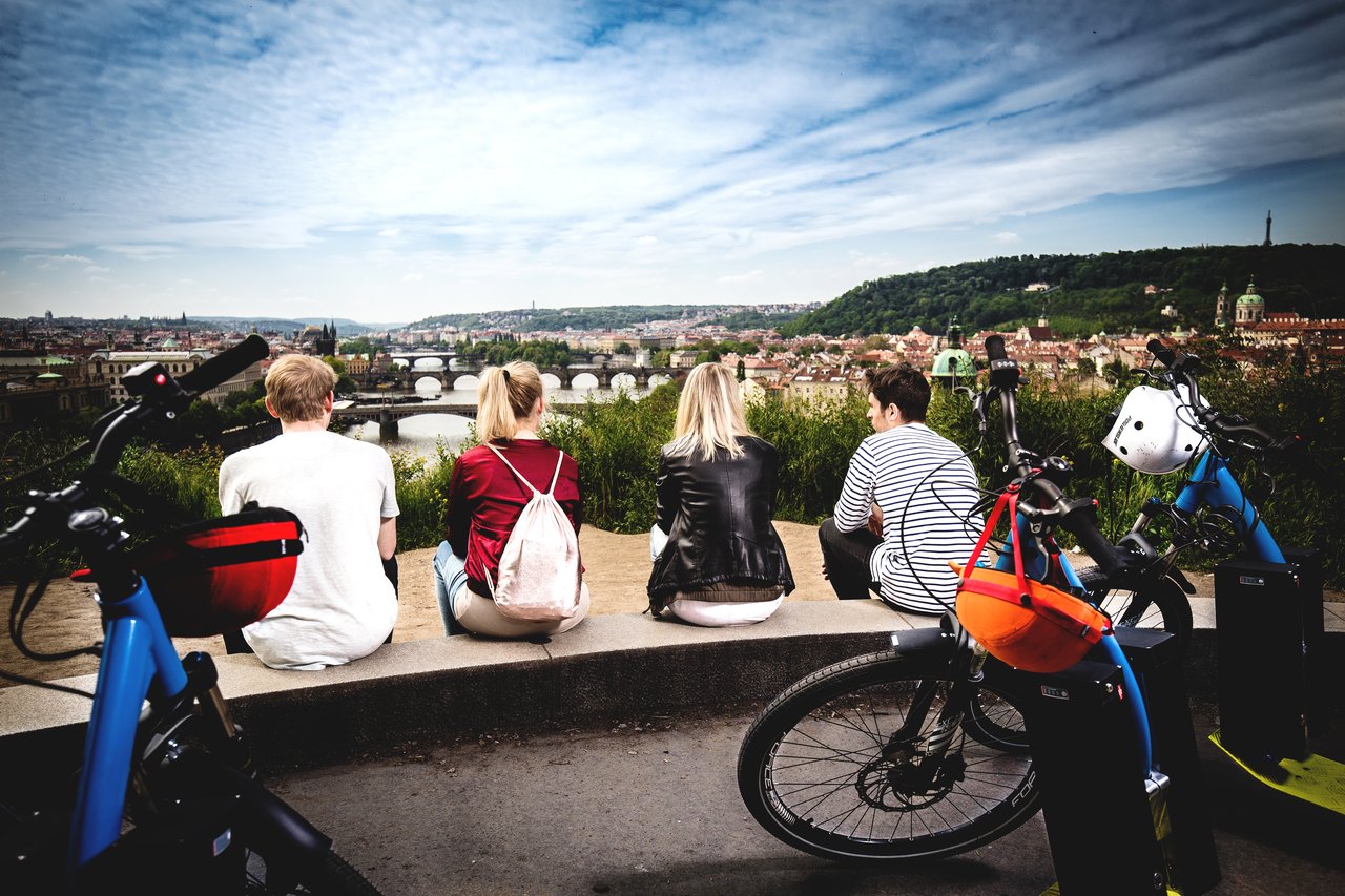 Quatre personnes assises dos à la caméra, contemplant la ville de Prague, avec des trottinettes électriques derrière elles