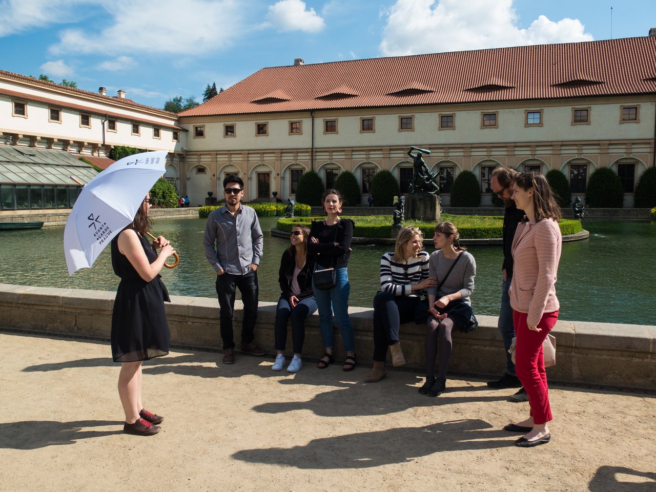 Un guide touristique, un parapluie à la main, s'adresse à sept personnes assis devant une fontaine sous un ciel ensoleillé.
