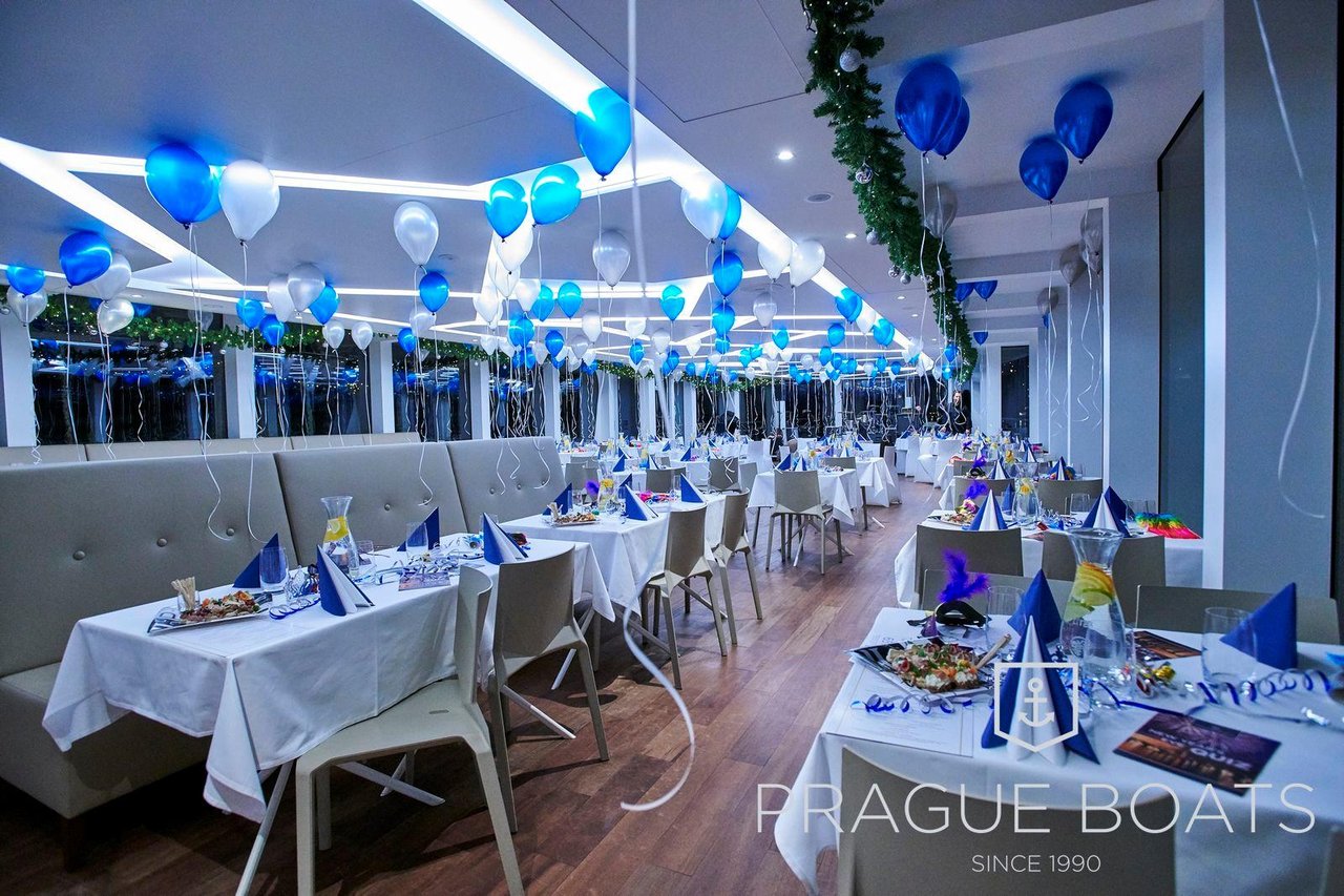 Plusieurs tables avec des fauteuils gris, la fête se passe sur un bateau il y a des ballons bleu et blanc