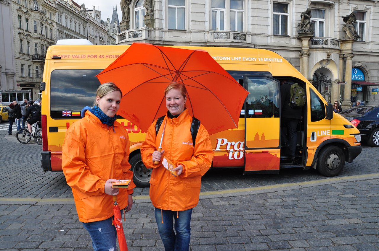 Deux femmes vêtues d'orange et munies de parapluies orange distribuent des prospectus pour le minibus.