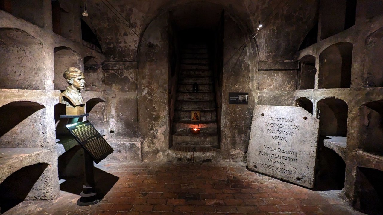 Une pièce souterraine faiblement éclairée, construite en pierre grise, contenant un buste sculpté reposant sur un piédestal.