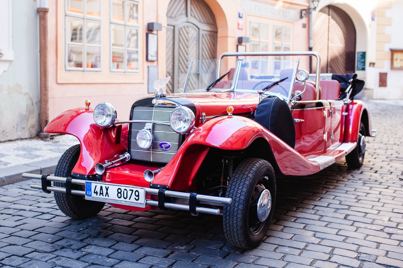 Une voiture historique rouge vif à ciel ouvert est garée sur une rue pavée devant des bâtiments historiques de Prague.