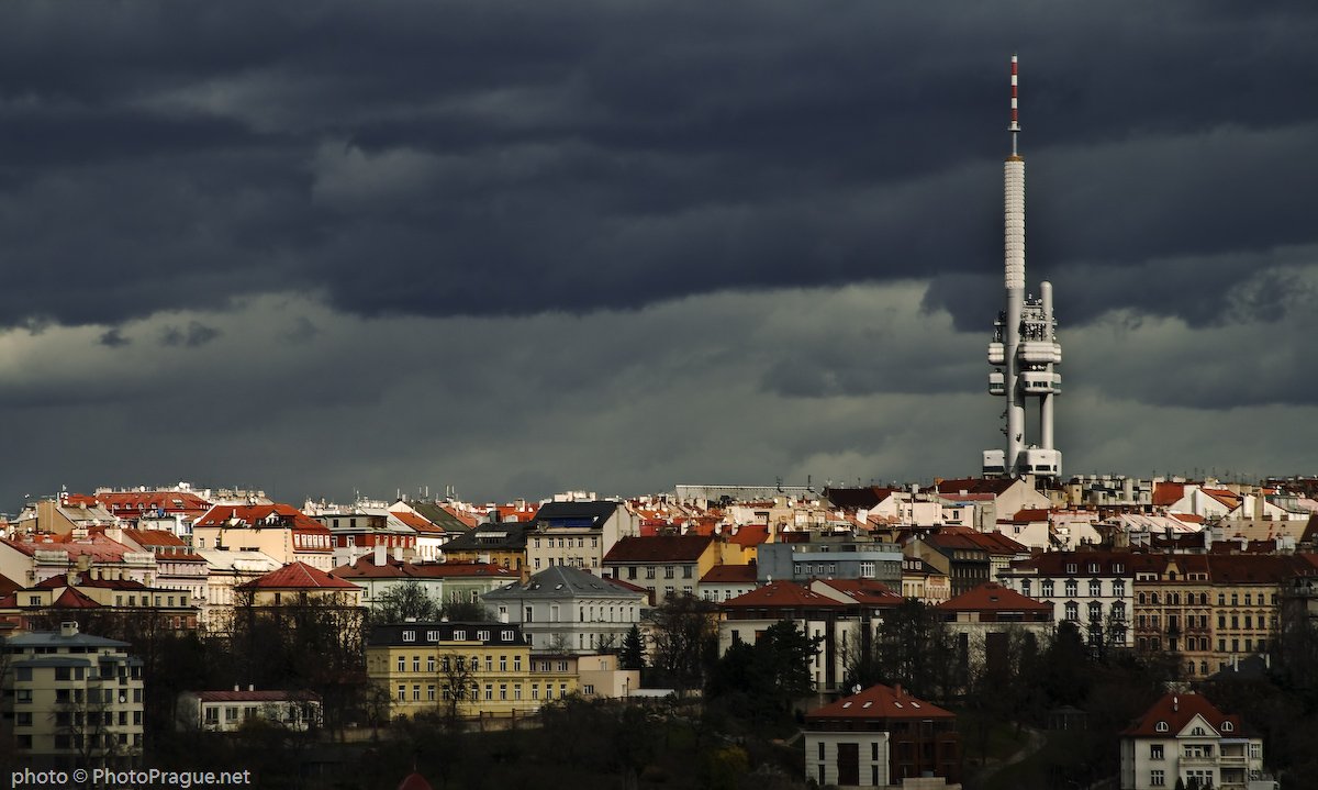 Une  tour blanche et pointue s'élève au-dessus d'une multitude de maisons avec un ciel très sombre de nuages gris.
