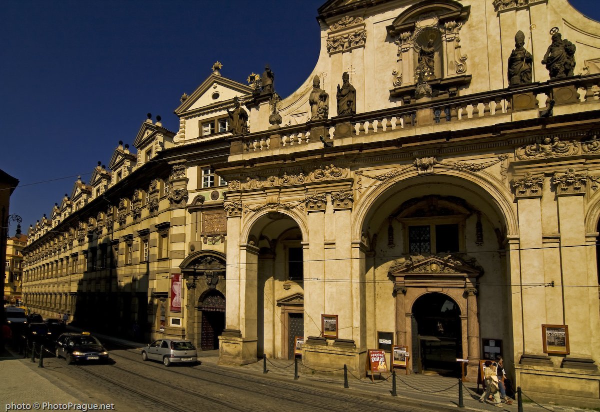 Façade en pierre sculptée d'un grand monument baroque , bordée par une rue pavée où circulent quelques voitures.