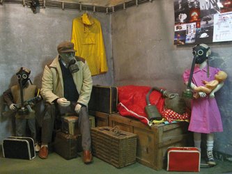 en:A family wearing gas masks in a concrete room where a poster depicting bombs is hanging ;fr:Une famille portant des masques à gaz dans une pièce en béton où est accrochée une affiche représentant des bombe