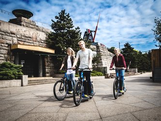 en:Three people on electric scooters in front of a stone building topped by a metronome ;fr:Trois personnes sur des trottinettes électriques devant un bâtiment en pierre surmonté d'un métronome