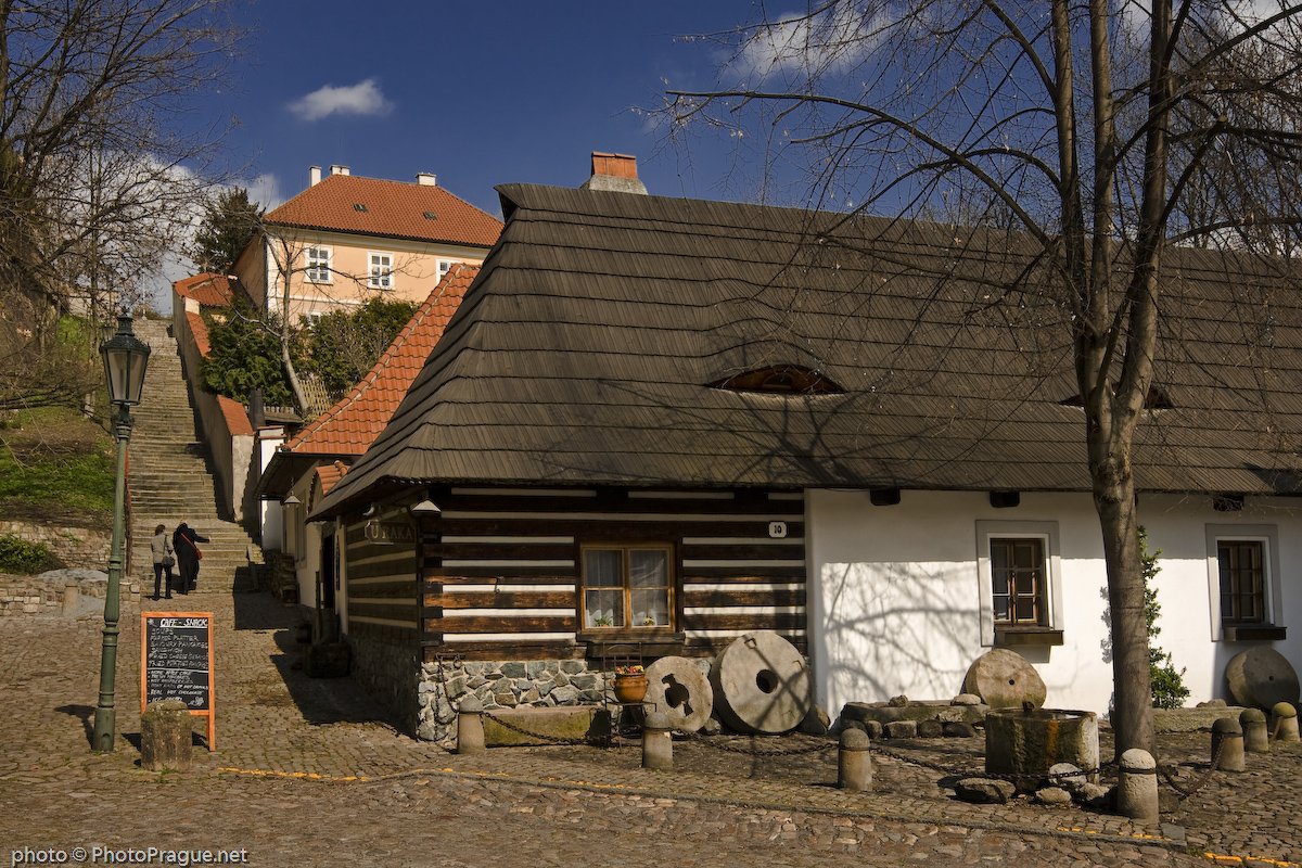 Une maison en bois chaleureuse se dresse au bord d'une ruelle pavée  sous un ciel bleu parsemé de nuages.
